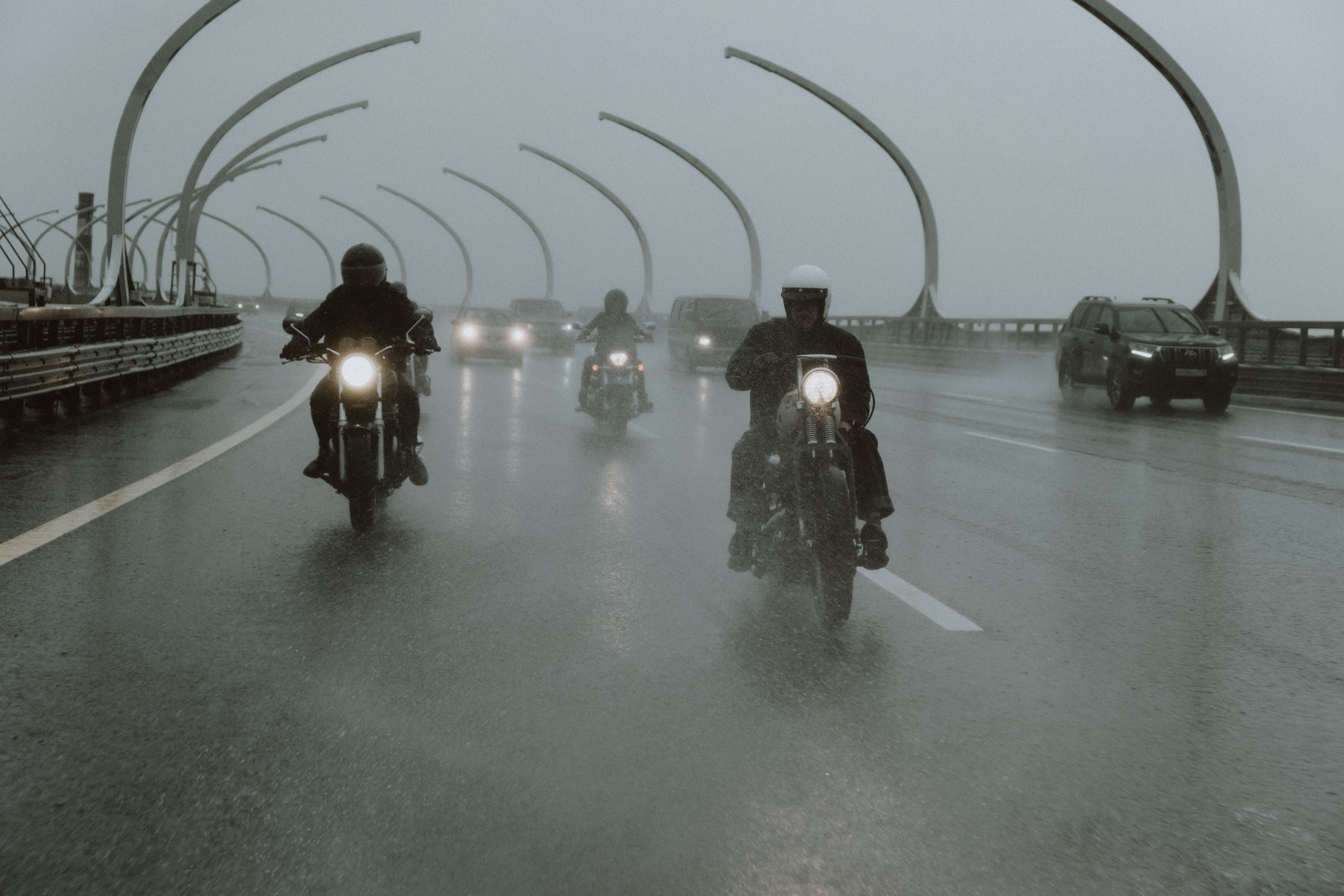 Motorcyclists riding across a wet highway bridge in the rain with headlights piercing the gloom